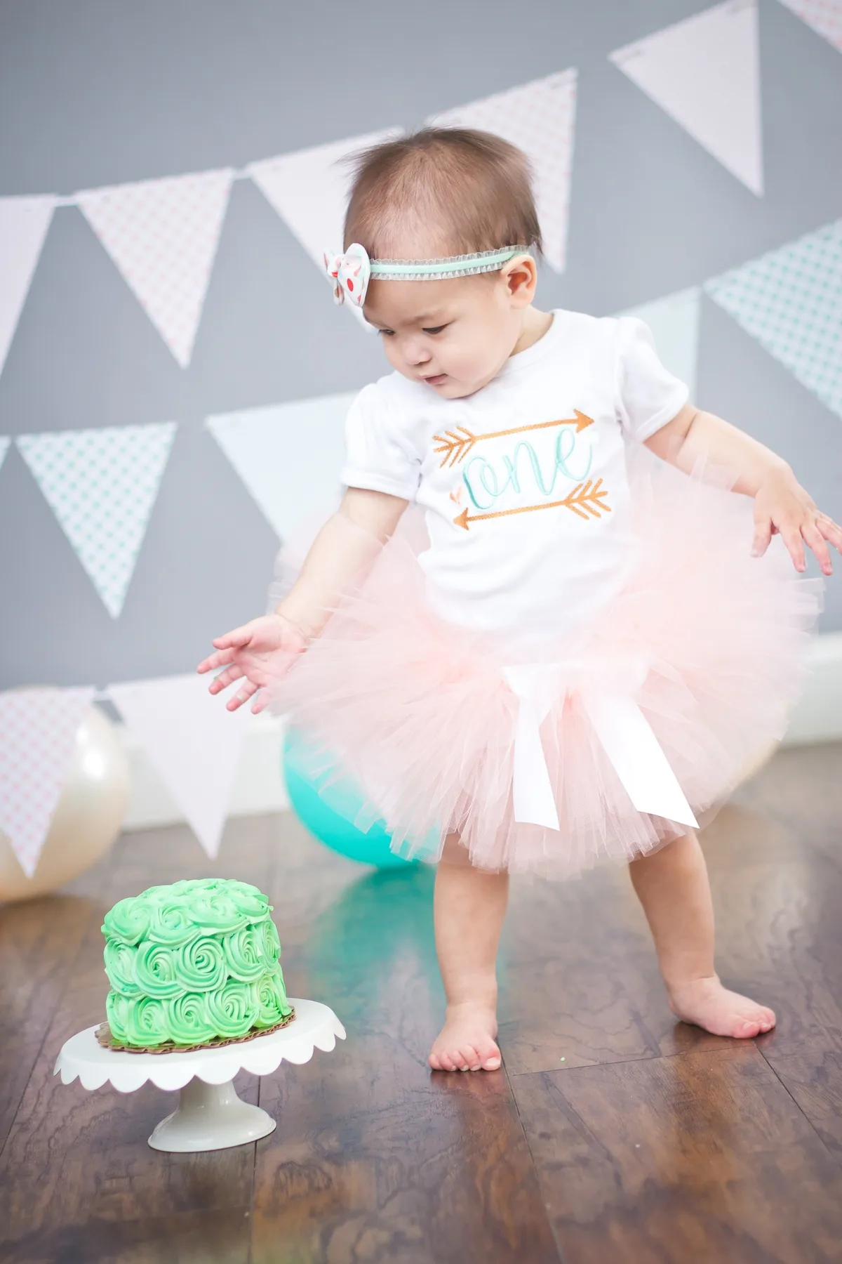 Baby girl in tutu and 'one' onesie standing next to a green rosette smash cake before the session begins