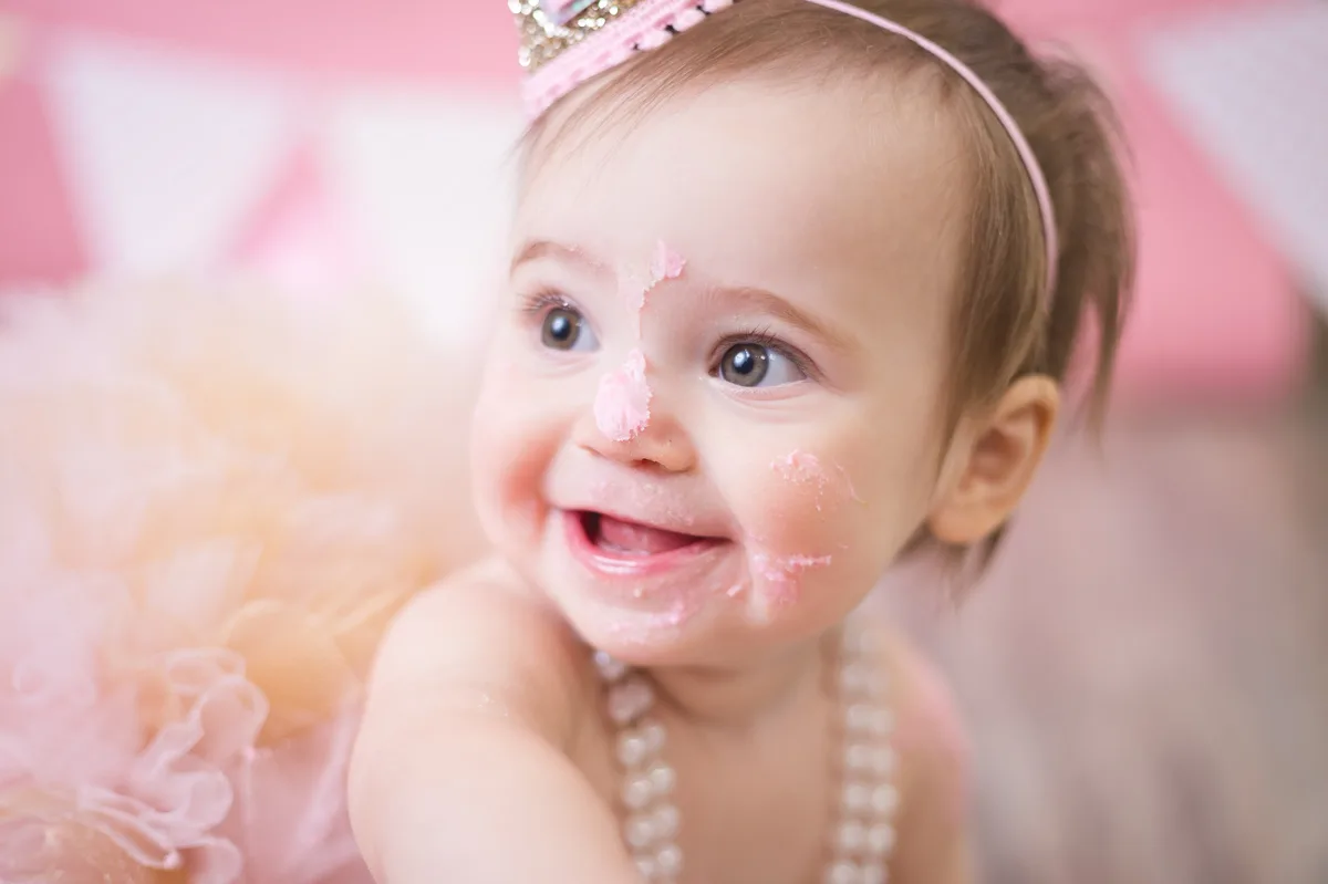 Baby girl with pink frosting on her nose and cheeks, laughing during cake smash session