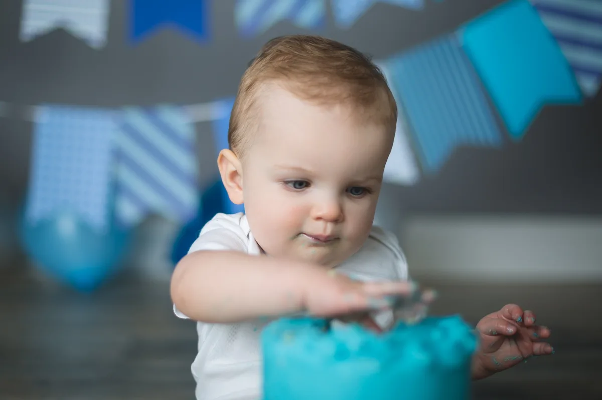 Baby reaching for blue birthday cake with blue banner and balloons