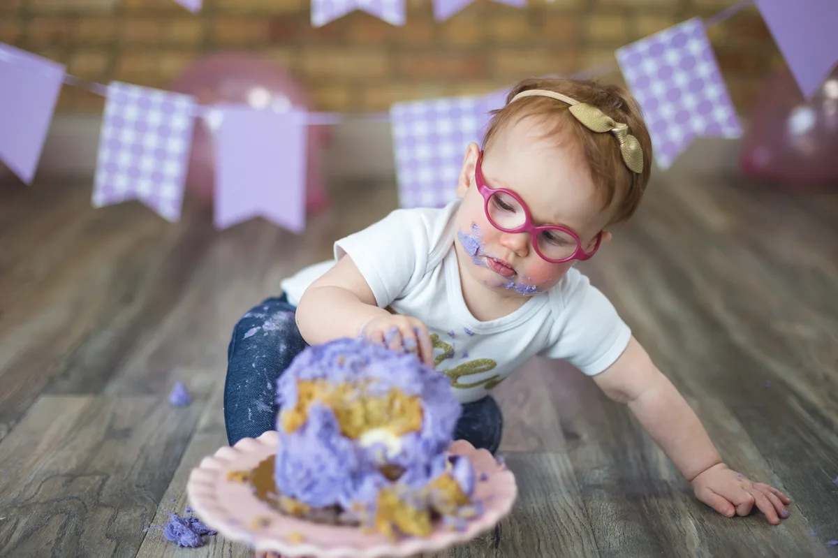 Baby girl with glasses digging into purple smash cake with purple banner